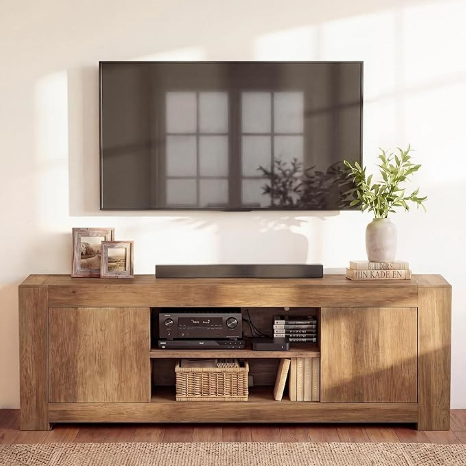 A rustic brown wooden TV stand media console with open shelving, styled with a soundbar, electronic equipment, books, and a vase of greenery, set against a warm off-white wall with a large framed TV above.
