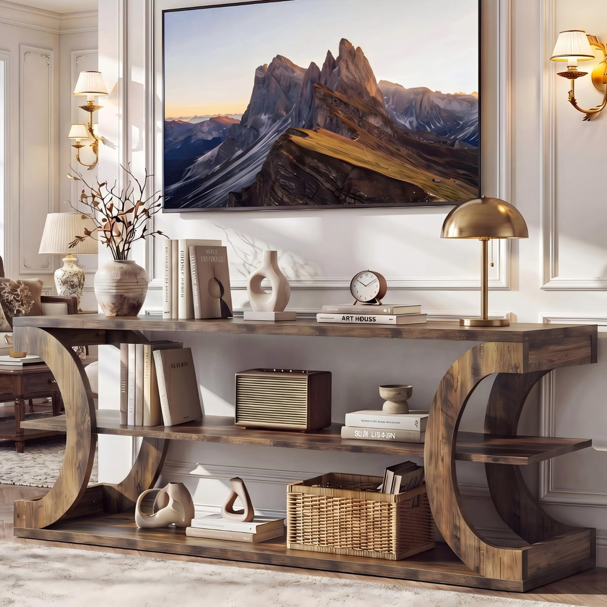 A rustic wooden console table with curved side supports and three shelves, styled with books, lamps, vases, and a basket in a bright living room. A large framed mountain landscape print hangs on the wall above.