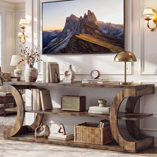 A rustic wooden console table with curved side supports and three shelves, styled with books, lamps, vases, and a basket in a bright living room. A large framed mountain landscape print hangs on the wall above.