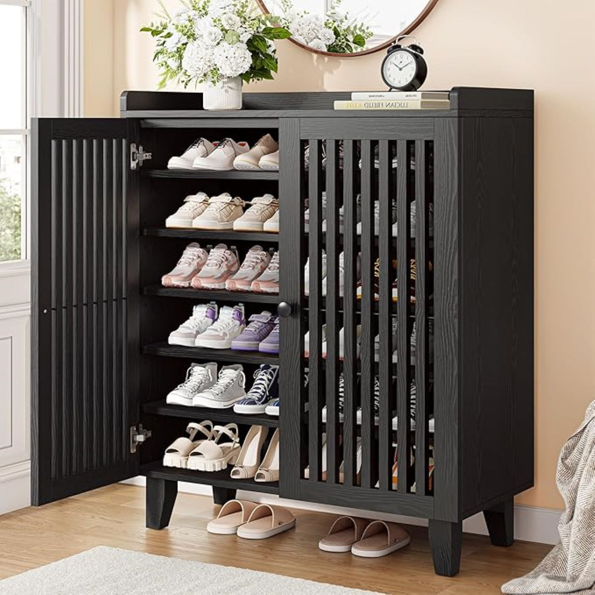 A tall black entryway shoe cabinet with one door open, showing multiple shelves filled with shoes. The cabinet has vertical slat detailing on the doors and stands on short legs in a styled room with a rug, floor, wall decor, and shoes placed outside the cabinet.
