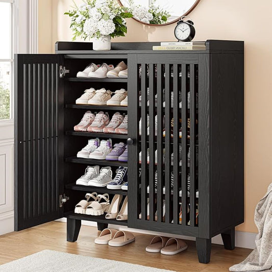 A tall black entryway shoe cabinet with one door open, showing multiple shelves filled with shoes. The cabinet has vertical slat detailing on the doors and stands on short legs in a styled room with a rug, floor, wall decor, and shoes placed outside the cabinet.