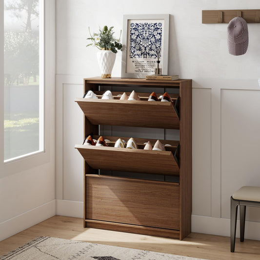 A tall wooden shoe cabinet with two open middle drawers and one lower drawer, styled in a bright entryway with shoes stored inside. A framed print, vase with greenery, and a small bench are visible around it.